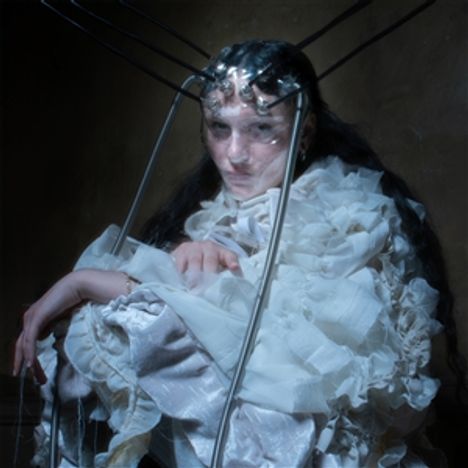 Es gibt keinen Text oder Logo. Eine Person in einem weißen, voluminösen Kleid trägt eine Art metallischen Kopfschmuck.