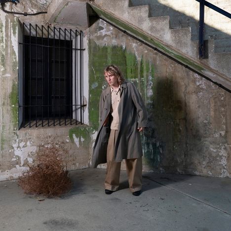 Ein Mensch steht in einem Hof vor einer verwitterten Wand mit Grünspan, neben einem Fenster mit Gitterstäben und einer Treppe.