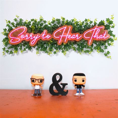 „Sorry to Hear That“ in pinkem Neonschriftzug auf Blättern. Zwei Figuren und ein „&“-Zeichen auf orangem Tisch.