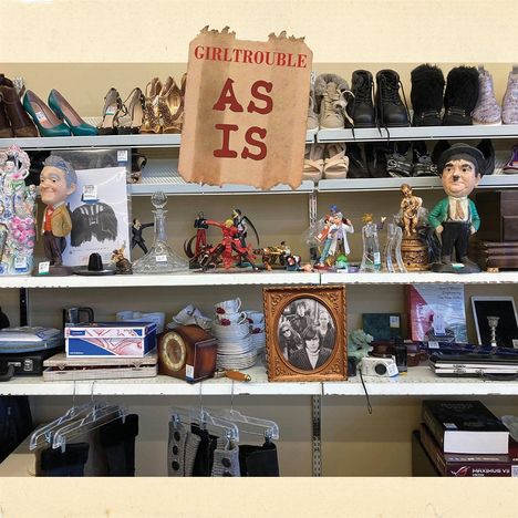 Text: "GIRLTROUBLE AS IS".   
Regal mit Schuhen, Dekorationen und Haushaltswaren.