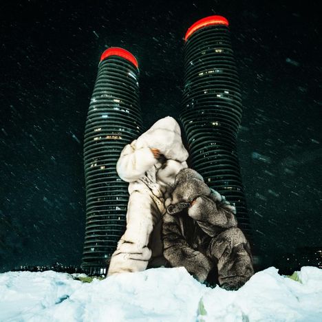 Zwei Personen in weißen und grauen Wintermänteln stehen vor beleuchteten, verdrehten Türmen im Schneesturm.