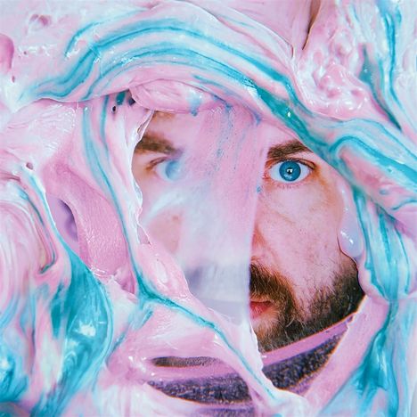 Ein Gesicht schaut durch eine Schicht aus rosa und blauem cremigen Material. Ein Auge ist klar sichtbar.