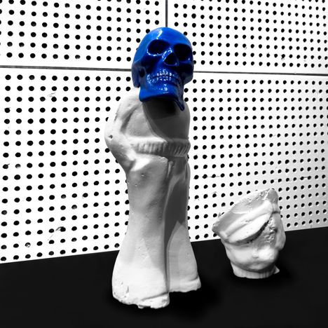 Eine abstrakte Skulptur mit einem blauen Totenkopf auf einem weißen Körper, neben einer ähnlichen Form auf schwarzem Boden.