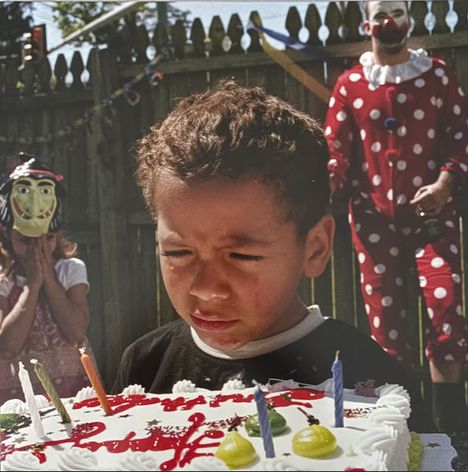 Ein Junge schaut auf einen Geburtstagskuchen mit bunten Kerzen. Hinter ihm steht eine Person in einem Clownskostüm.