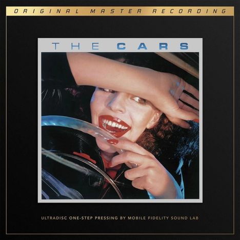 Text: "THE CARS". Eine lachende Frau hinter einem Lenkrad mit rotem Lippenstift und lackierten Nägeln.