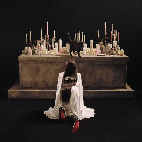 Rückenansicht einer Frau im weißen Kleid mit "ESTRUS" darauf, kniend vor einem Altar mit vielen Kerzen und einer schwarzen Katze.