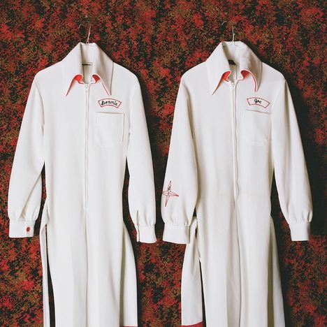 Links "Bonnie", rechts "Joe". Zwei weiße Overalls mit roten Details vor einem gemusterten Hintergrund.