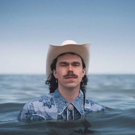 Mann mit Cowboyhut und Schnurrbart steht im Wasser, trägt ein blaues Hemd. Im Hintergrund ist der Horizont sichtbar.