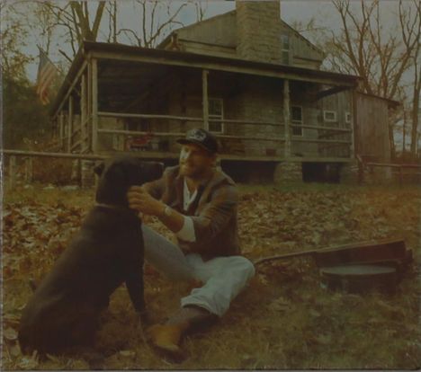 Eine Person mit Kappe streichelt einen schwarzen Hund vor einem rustikalen Haus; eine Gitarre liegt im Gras.