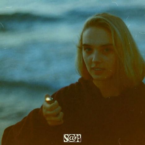 Eine Person hält ein Feuerzeug am Strand. Im Hintergrund verschwommenes Wasser. Unten ist ein "S@♈" Logo.