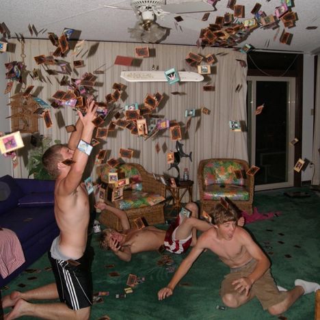Drei junge Männer werfen spielend Karten in die Luft in einem Wohnzimmer mit bunten Möbeln und Teppich.