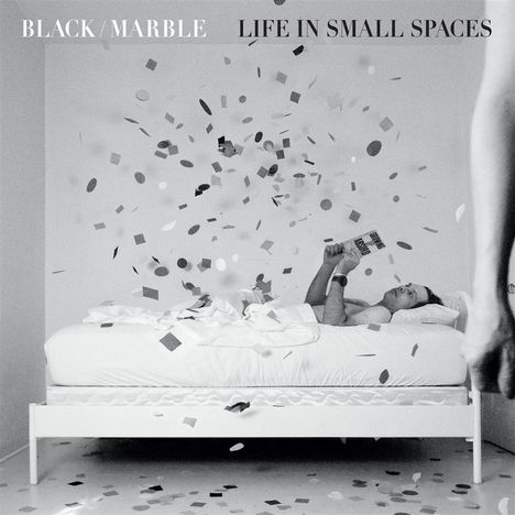 "BLACK/MARBLE LIFE IN SMALL SPACES" Oben, Konfetti wirbelt um liegende Person mit Buch auf Bett.