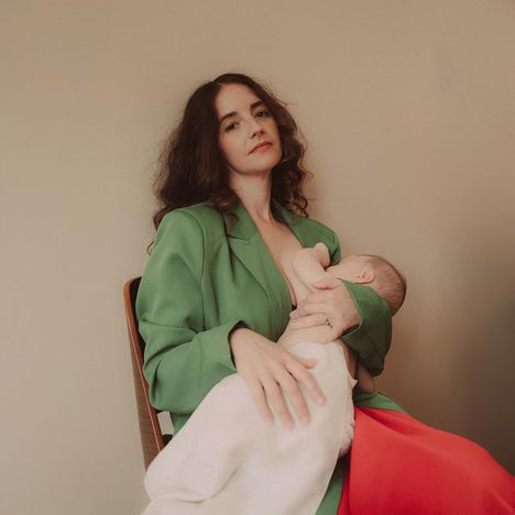 Eine Frau in einem grünen Blazer stillt ein Baby und sitzt auf einem Stuhl. Das Baby ist in ein weißes Tuch gehüllt.