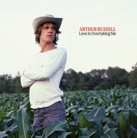 „ARTHUR RUSSELL Love Is Overtaking Me“. Eine Person mit Hut steht in einem Feld, umgeben von Pflanzen.