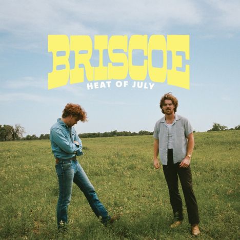 "BRISCOE HEAT OF JULY" in gelber Schrift. Zwei Personen stehen auf einer grünen Wiese unter einem blauen Himmel.