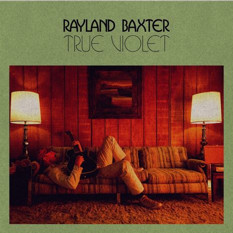 "Rayland Baxter. True Violet." Eine gemütliche Szenerie mit Mann und Gitarre auf einem Sofa zwischen zwei Lampen.