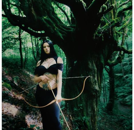 Ich kann den Text nicht identifizieren. Eine Frau mit Pfeil und Bogen in einem grünen Wald, getragenes Kleid schwarz.