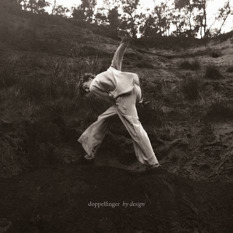"doppelfinger by design" – Eine Person in heller Kleidung bewegt sich in dramatischer Pose auf einem Hügel mit Vegetation.