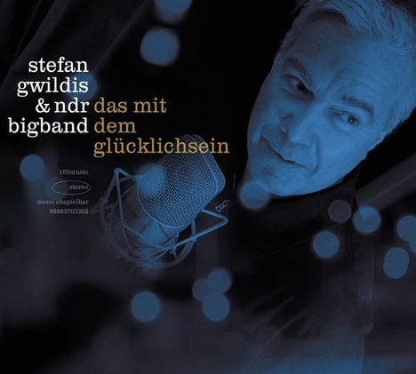 "stefan gwildis & ndr bigband das mit dem glücklichsein". Ein Mann mit Mikrofon, blaue Tönung, Bokeh-Effekte.
