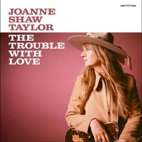 Text: "JOANNE SHAW TAYLOR, THE TROUBLE WITH LOVE." Eine Frau in Hut und Jacke hält eine Gitarre vor rosa Hintergrund.