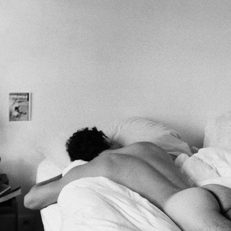 Ein schwarz-weißes Foto zeigt eine Person, nackt auf dem Bauch in einem Bett liegend, in einem spartanischen Raum mit einem Poster an der Wand.