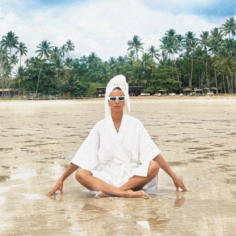 Keine erkennbaren Texte. Eine Person in weißem Bademantel sitzt am Strand im Lotussitz, Palmen im Hintergrund.