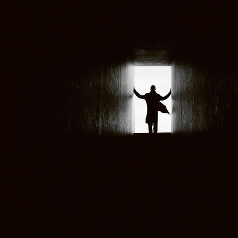 Ein Mann im Mantel steht in einem dunklen Raum vor hell beleuchtetem Ausgang, Arme ausgestreckt an den Wänden.