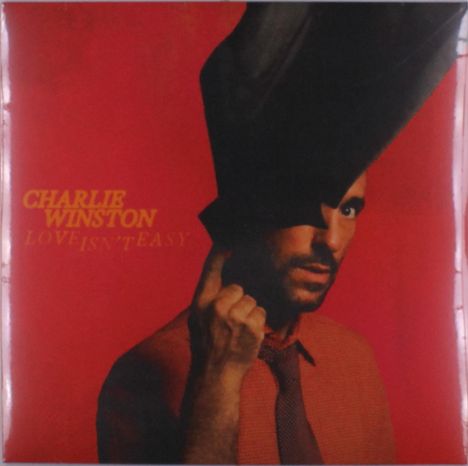 Text: "Charlie Winston, Love Isn't Easy." Ein Mann mit Hut auf rotem Hintergrund, stilvolle Pose.