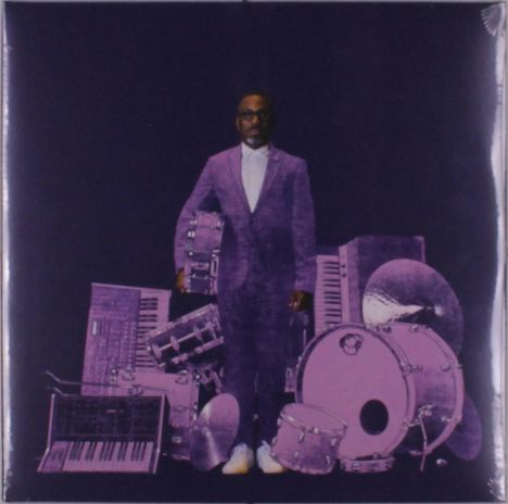 Ein Mann in einem Anzug steht umgeben von lila Instrumenten wie Schlagzeug und Keyboards vor einem dunklen Hintergrund.