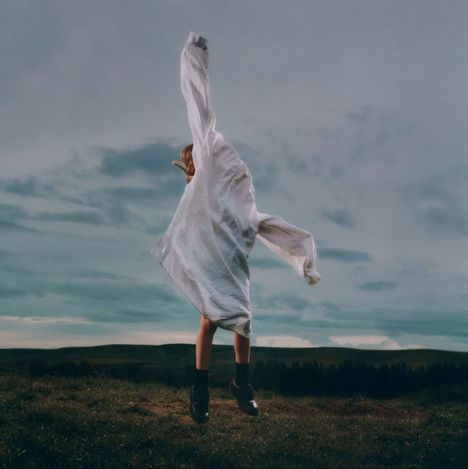 Keine Texte sichtbar. Eine Person in einem weißen Mantel springt auf einer Wiese vor einem bewölkten Himmel.