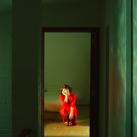 Es gibt keinen Text. Eine Frau in einem roten Kleid kauert nachdenklich in einem beleuchteten Türrahmen.