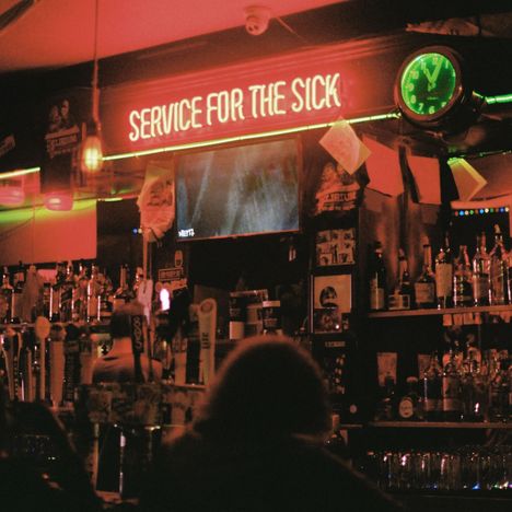 Der Text "SERVICE FOR THE SICK" leuchtet in Neon. Eine Bar mit vielen Flaschen, Zapfhähnen und eine grün beleuchtete Uhr.