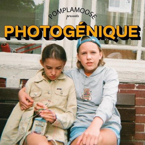 "POMPLAMOOSE presents PHOTOGÉNIQUE." Zwei Kinder sitzen auf einer Bank, eine umarmt die andere, beide schauen entspannt.