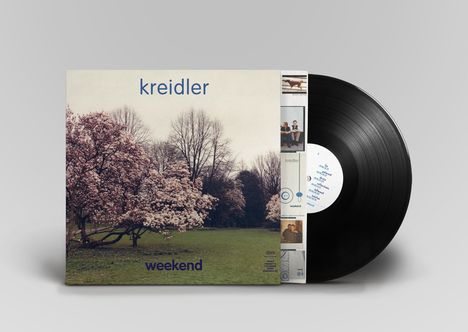 "kreidler" oben, "weekend" unten. Ein blühender Baum mit rosa Blüten vor einer Wiese und kahlen Bäumen im Hintergrund.