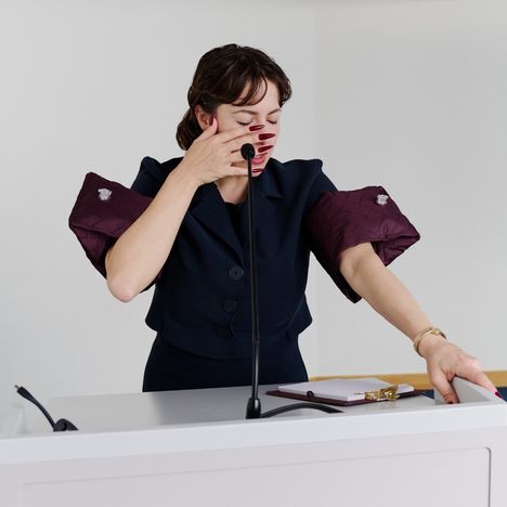 Eine Frau mit geschlossenen Augen steht an einem Rednerpult, trägt ein dunkles Outfit und hat lange rote Fingernägel.