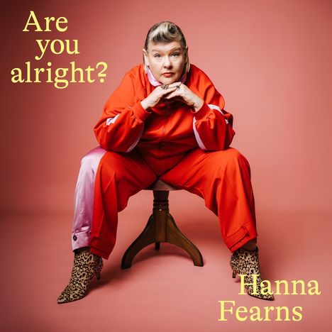 Text: "Are you alright?" Eine Person im roten Anzug mit Leopardenmuster-Schuhen sitzt auf einem Hocker vor rosa Hintergrund.