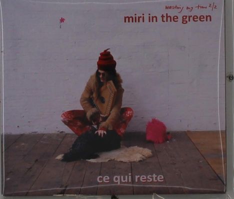 „miri in the green“, „ce qui reste“: Frau in bunter Kleidung hockt auf Holzboden, streichelt Hund.