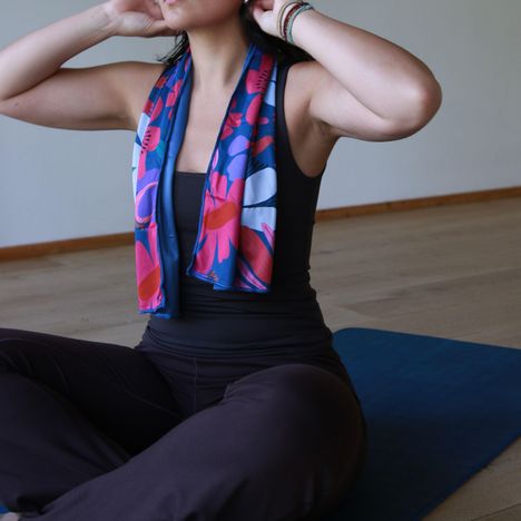 Frau auf blauer Matte in Yoga-Pose, trägt ein farbenfrohes Tuch um den Hals und dunkle Yoga-Kleidung.