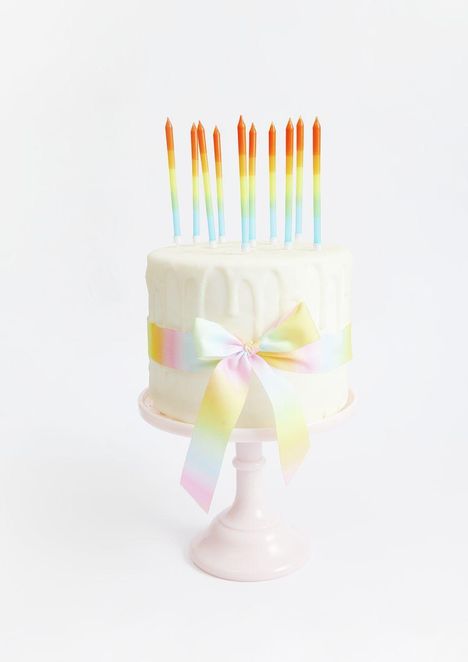 Weiße Torte mit buntem Band und zwölf bunten Kerzen auf pastellfarbenem Kuchenständer.