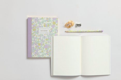Notizbuch mit floralem Muster, Text "NOTEBOOK". Geöffnete, punktierte Seiten. Stift, Anspitzer, Holzspäne daneben.