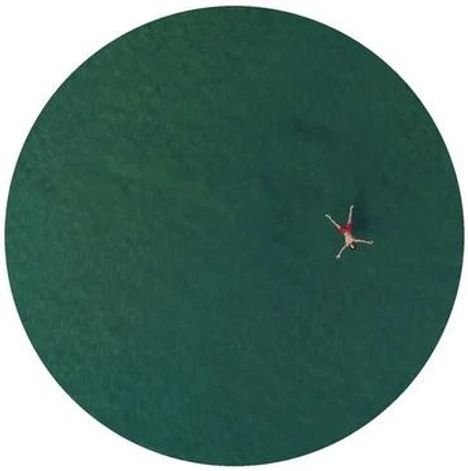 Keine Texte sichtbar. Obenansicht eines Menschen in roter Kleidung schwimmend im weiten, grünen Wasser. Rundes Format.