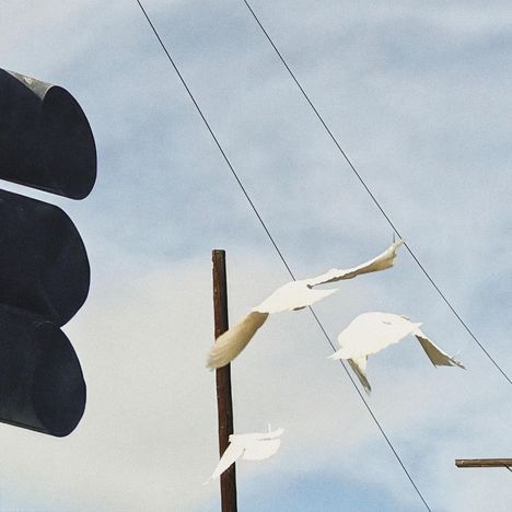 Drei weiße Tauben fliegen vor wolkigem Himmel. Links ein dunkles Ampelgehäuse, dahinter Telefonleitungen.