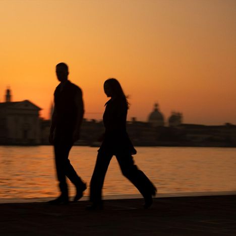 Zwei Personen im Schatten laufen vor einem orange-goldenen Sonnenuntergang entlang eines Gewässers mit Stadt-Silhouette.