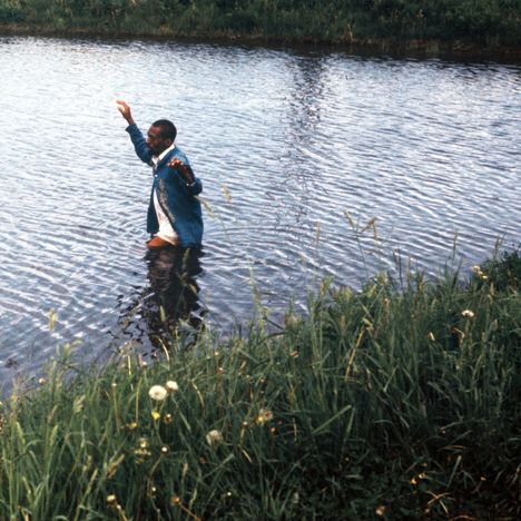 Ein Mann watet durch einen flachen, klaren Fluss, umgeben von grüner Vegetation am Ufer.
