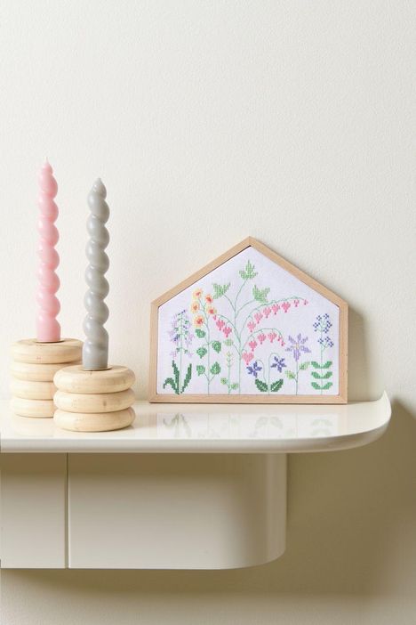 Stickbild mit bunten Blumen in Hausform neben zwei gedrehten Kerzen, eine rosa und eine graue, auf Holzsockeln.