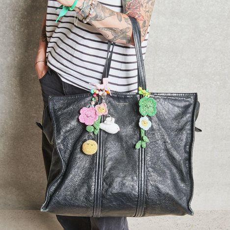 Ein großer schwarzer Lederbeutel mit buntem gehäkeltem Blumen- und Tieranhänger, gehalten von tätowiertem Arm in gestreiftem Shirt.