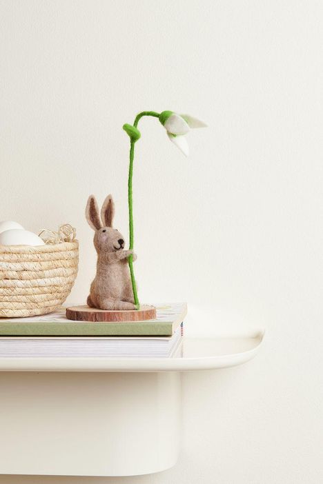 Ein Filzhase hält einen Schneeglöckchen-Stab. Daneben ein Korb mit Eiern auf Büchern, alles auf einem Regal.