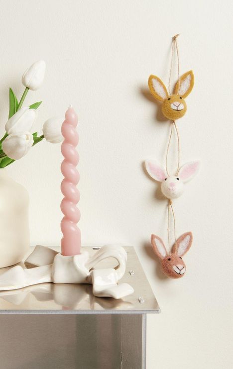 Kein Text. Weiße Tulpen in Vase, rosa gedrehte Kerze im geschwungenen Halter, Filzhäschen als Deko hängend.