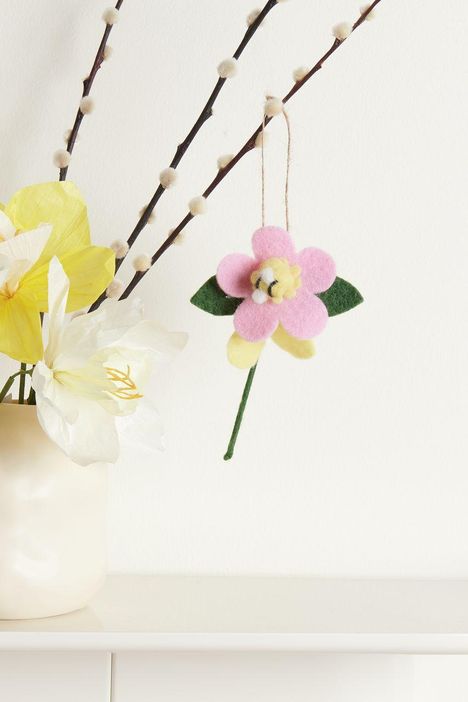 An einem kahlen Zweig hängt ein rosa Filzblume. Daneben gelbe und weiße Blumen in einer Vase.