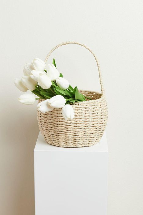 Ein heller Korb voller weißer Tulpen, stehend auf einem weißen Sockel vor einem neutralen Hintergrund.